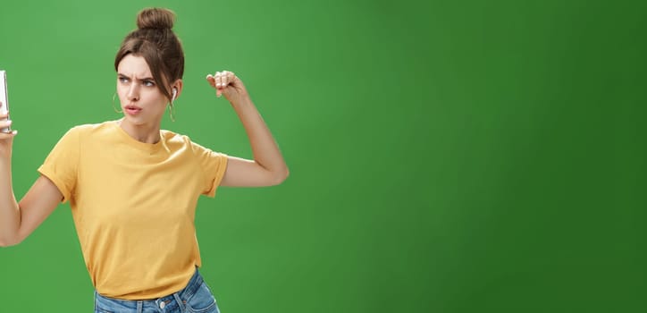 Woman making serious look to take selfie while listening music in wireless earphones posing and staring at smartphone screen daring and aggressive, liking post photos online over green background. Technology and lifestyle concept