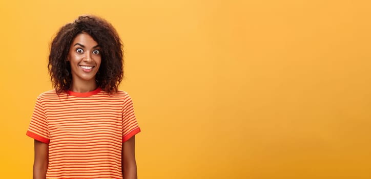 Waist-up shot of amazed and excited charming african american woman with curly hairstyle popping eyes from thrill and joy smiling broadly being surprised by great gift over orange background. Lifestyle.