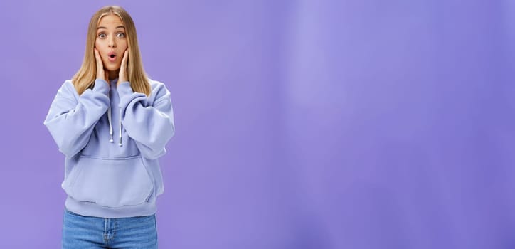 Indoor shot of amused and thrilled surprised good-looking young woman in stylish hoodie folding lips in wow sound popping eyes at camera shocked and excited learning amazing news over purple wall. Lifestyle.