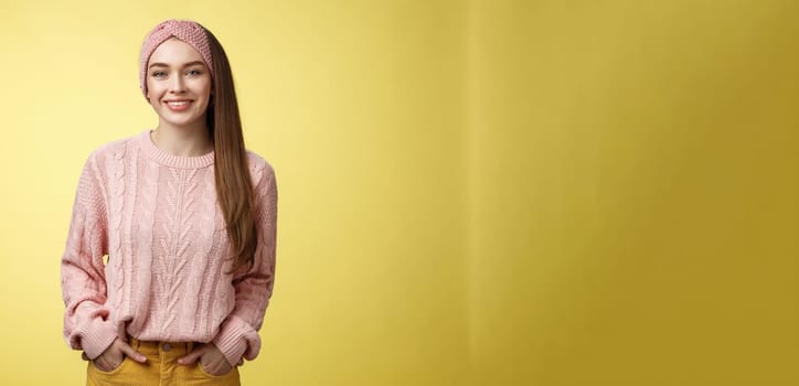 Charming friendly young girl in knitted sweater and headband holding hands in pockets, smiling, looking positive motivated achieving goals having great day, posing positive against yellow background.