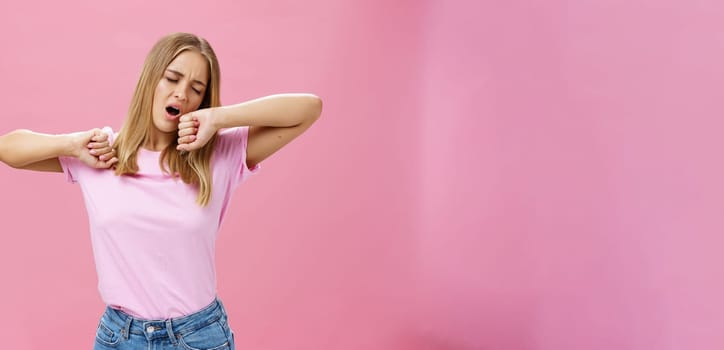 Too tired to work today. Lazy and exhausted attractive young female student doing homework all night yawning with closed eyes while stretching hands from after tiresome project over pink wall. Body language concept
