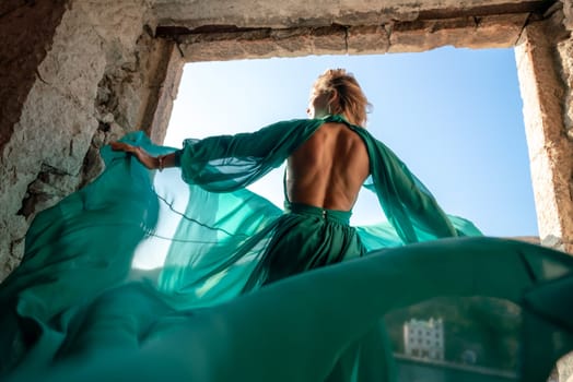 Rear view of a happy blonde woman in a long mint dress posing against the backdrop of the sea in an old building with columns. Girl in nature against the blue sky