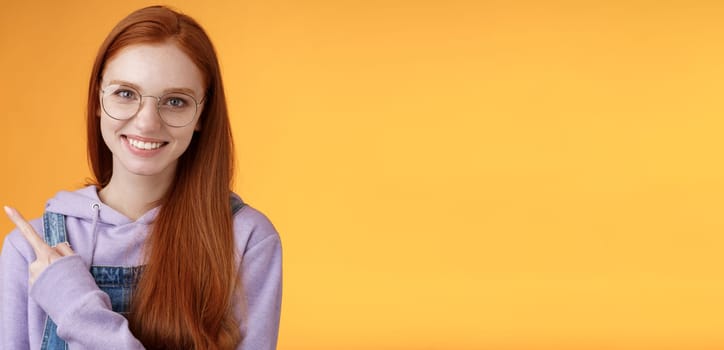 Friendly good-looking modern redhead young girl pointing left index finger showing awesome place suggesting go hang out smiling joyfully casually talking discuss new product, orange background.