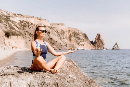 Yoga on the beach. A happy woman meditating in a yoga pose on the beach, surrounded by the ocean and rock mountains, promoting a healthy lifestyle outdoors in nature, and inspiring fitness concept