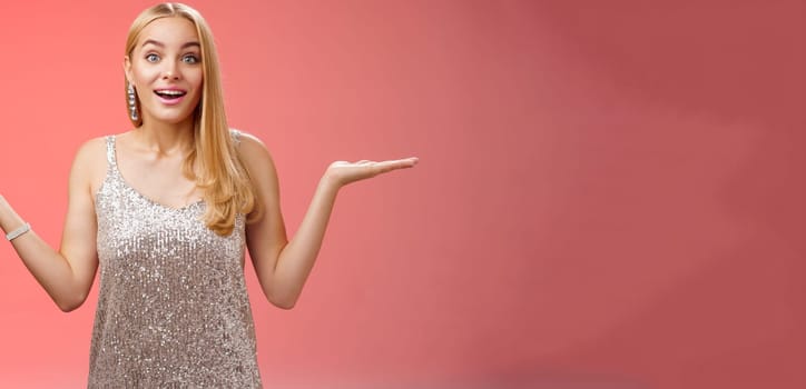 Carefree fascinated young blond girl 20s in silver glittering evening party dress raising hands sideways weighing shrugging smiling amused search right choice, deciding what do red background.