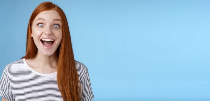 Surprised pleased happy impressed redhead european girl 20s reacting amused wide eyes look admiration joy receive incredible offer standing excited express thrill upbeat feelings, blue background.