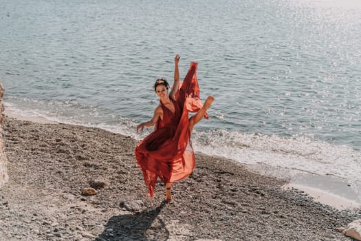 Woman red dress sea. Female dancer in a long red dress posing on a beach with rocks on sunny day. Girl on the nature on blue sky background