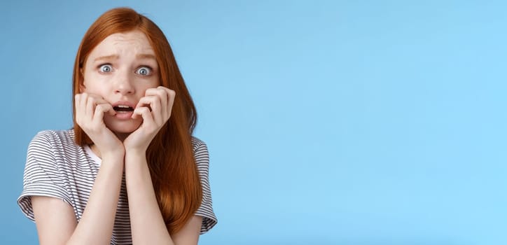 Scared speechless stunned frightened redhead girl trembling fear wide eyes terrified biting fingernails frowning shaking anxiously standing blue background gasping shocked. Emotions concept