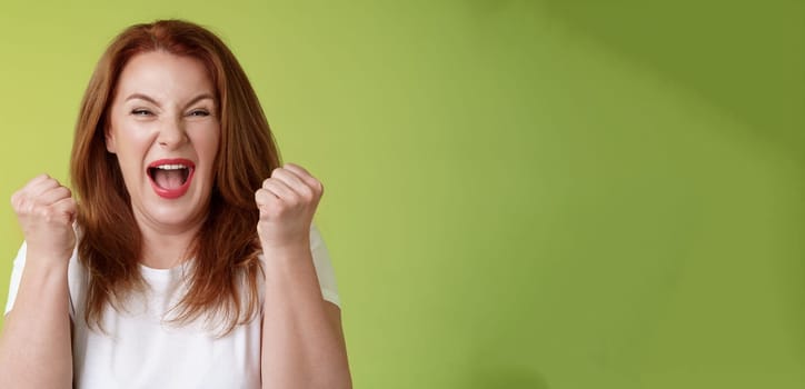 Yeah we did it. Joyful lucky redhead middle-aged female winner pump fists up celebration success gesture yelling triumph joy smiling broadly celebrate awesome news stand green background happy.