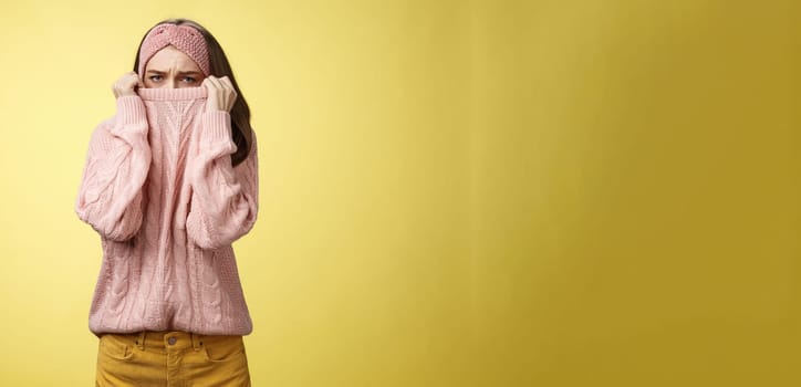 Sad silly, gloomy cute young girl hiding face in sweater collar pulling clothes on nose frowning displeased, unhappy, looking indignant and reluctant standing disappointed over yellow background.