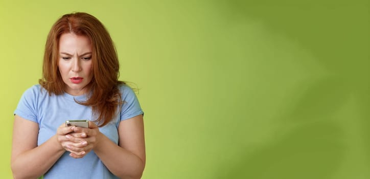 Lifestyle Concept - Confused unsure redhead middle-aged woman learn how use social media. trying understand emoji look intense focused smartphone display reading important news message stand green background.