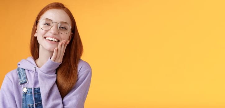 Charismatic happy tender redhead smiling european girl 20s glasses hoodie denim overalls having fun enjoying summer holidays chilly evening laughing joyfully touch pure clear skin, orange background.