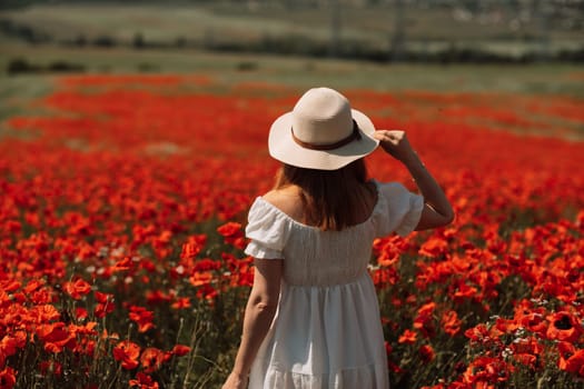 Field of poppies woman. Happy woman in a wight dress and hat stand through a blooming field of poppy. Standing with her back raised her hands up. Field of blooming poppies