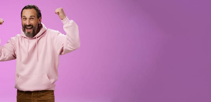 Yeah who cares age. Carefree delighted happy old man bearded in trendy pink hoodie raise fists joyfully triumphing having fun glad win celebrating success accomplish goal, posing purple background.