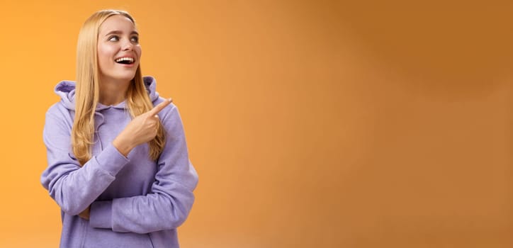 Amused carefree joyful blond european woman in hoodie pointing looking right pleased smiling happily enjoying interesting fascinating peromance making choice in store shopping, orange background.