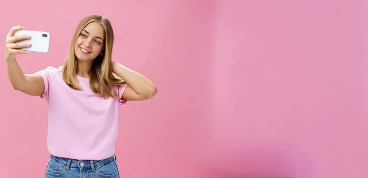 Female beauty blogger taking selfie post new look online. Portrait of charming tanned young woman in t-shirt touching hair gently pulling hand with smartphone near face taking photo over pink wall. Technology concept