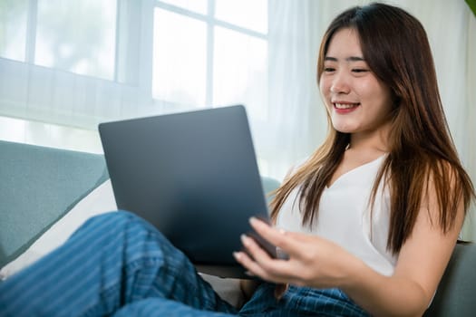 Happy woman typing email on notebook computer, Asian young female smiling sitting relaxing on sofa using laptop in living room at home, freelance browsing through the internet during free time