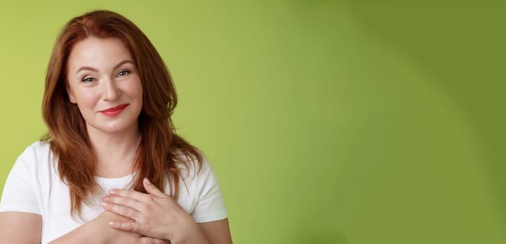 Lovely pleased tender redhead middle-aged ginger woman. press hands heart feel touched grateful appreciate sincere heartwarming moment admire thankfully grin look camera glad green background.