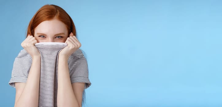 Silly flirty amused attractive playful redhead girlfriend hiding face pulling t-shirt head squinting devious mysteriously giggle laughing hope disguise pranking friend standing blue background.