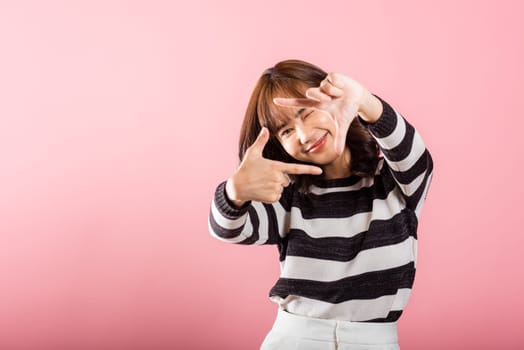 A cheerful Asian photographer frames her vision with a finger, expressing the concept of photography. Studio shot on pink background, highlighting inspiration and creativity.