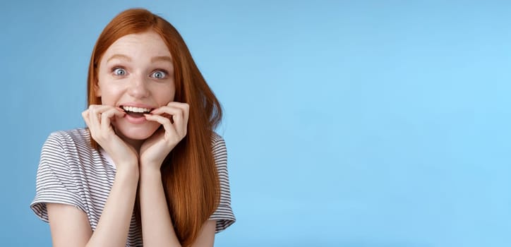 Excited amazed attractive redhead girl blue eyes look fascinated affection smiling desire bite fingernails eager feel astonished cannot wait bite tasty food, standing blue background thrilled.