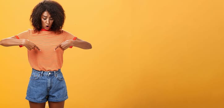 Portrait of worried questioned and surprised african american female with curly hairstyle looking and pointing at stomach or belly feeling discomfort and problems with health over orange background. Lifestyle.