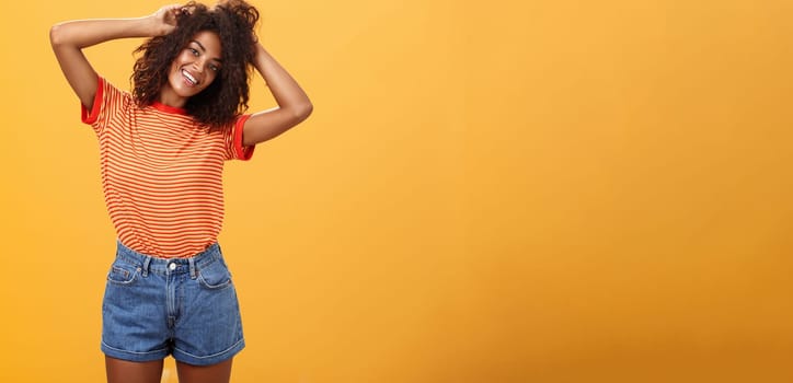 Time start living life fullest. Joyful optimistic woman having fun during vacation tilting head touching curly hair and enjoying summer sunshine in trendy striped t-shirt and shorts over orange wall.