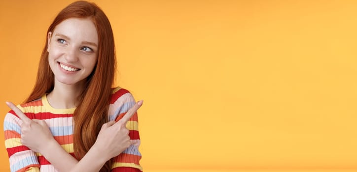 Dreamy cute redhead girl planning were go summer holidays have different choices picking variant look intrigued smiling pleased pointing sideways left right taking decision, orange background.