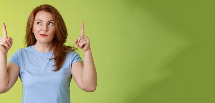 Curious thoughtful cute redhead female middle-aged ginger mother smirking thinking look aside pointing up index fingers deciding choice pondering choosing between variants green background.