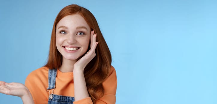 Cute tender chatismatic glad smiling redhead girl presenting awesome product show object palm hold hand raised blank copy space grinning impressed receive silly charming gift, blue background.