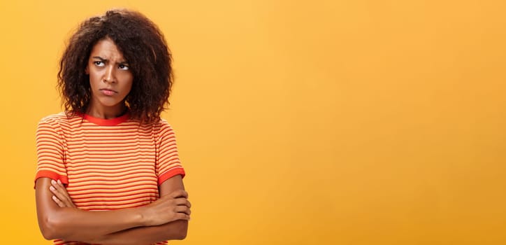 Why life so cruel and unfair. Gloomy displeased and sad cute african american young woman with curly hairstyle crossing arms on chest in offended pose frowning looking left jealous and unhappy.