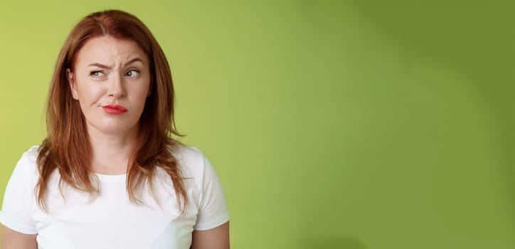 Perplexed doubtful unsure redhead wife smirking unsatisfied frowning look aside displeased disappointed hesitating pondering strange advice thoughtfully stare away stand green background.