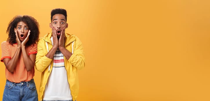 Portrait of shocked and stunned speechless girlfriend dropping jaw from amazement with boyfriend feeling amazed from shook news posing together surprised and astonished over orange wall. Lifestyle.