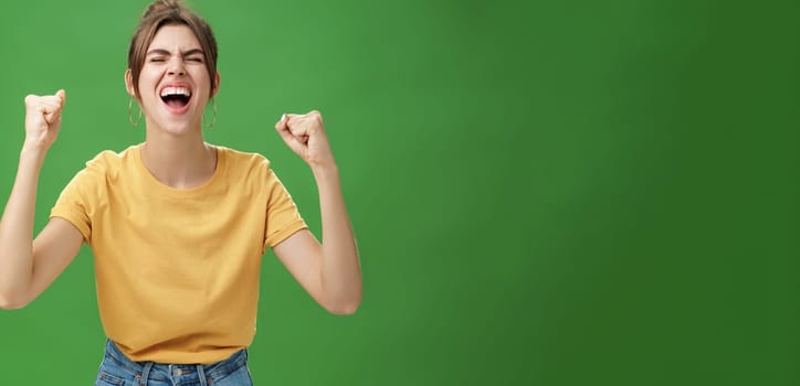 Waist-up shot of charismatic energized and excited female in yellow t-shirt closing eyes yelling from joy and happiness raising hands in cheer celebrating successful news over green background. Lifestyle.