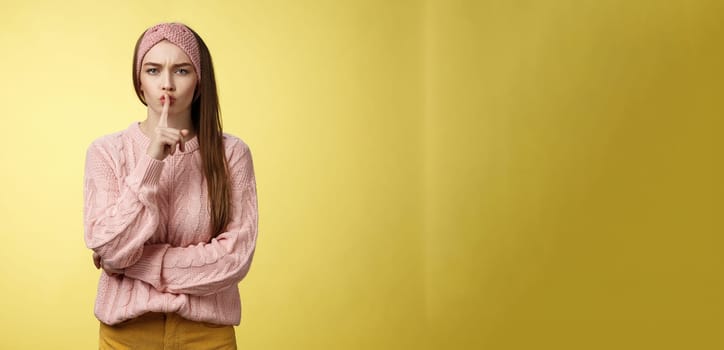 Keep mouth shut. Serious-looking bossy attractive young 20s woman in sweater, headband shushing making shhh gesture holding index finger on lips, gossiping, spread rumors over yellow background.