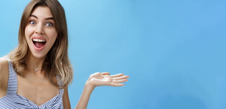 Waist-up shot of charismatic upbeat enthusiastic cute female blogger raising hand over copy space and holding camera as if recording video from vacation showing cool product over blue background. Lifestyle.