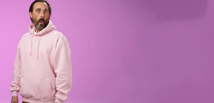 Impressed speechless shocked adult bearded man grey hair in cool pink hoodie folding lips wow popping eyes camera surprised astonished intrigued, standing curious amazed purple background.