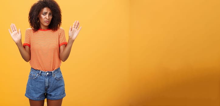 Silly insecure and sad dark-skinned female model in trendy striped t-shirt and shorts raising arms in surrender frowning being uninvolved and unaware standing clueless over orange background. Lifestyle.