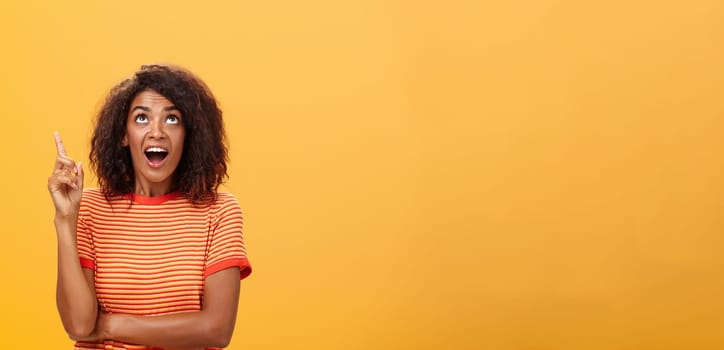 Waist-up shot of interested curious good-looking dark-skinned female in striped t-shirt talking asking question about curious star looking and pointing up with joy over orange background. Copy space