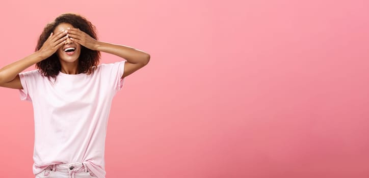Portrait of charming joyful dark-skinned playful woman with curly hair in t-shirt closing eyes and counting ten with broad happy smile playing hide n seek or waiting for surprise over pink background. Lifestyle.