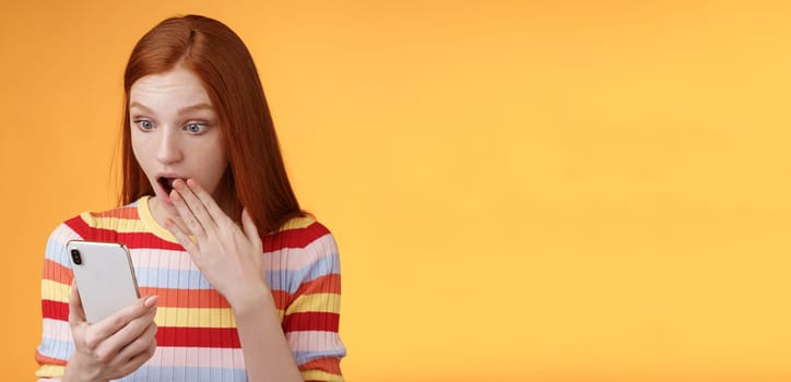 Lifestyle. Amazed speechless young teenage redhead girl student gasping drop jaw say omg wow cover opened mouth palm look shocked surprised smartphone display reading fresh gossips orange background.