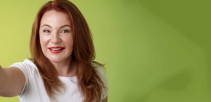 Close-up joyful enthusiastic redhead alluring middle-aged woman extand arm towards camera taking selfie smartphone smiling broadly posing photograph taking picture device green background.