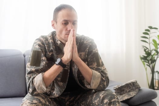 Portrait of calm serious Caucasian military man wearing camouflage uniform and cap sitting on a sofa, having depressed facial expression. High quality photo