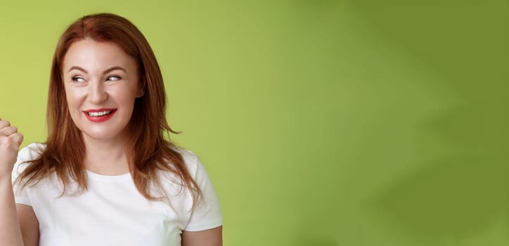 Intrigued charismatic redhead middle-aged woman smiling temtation interest pointing looking left curiously check-out cool promo share awesome place location stand green background.