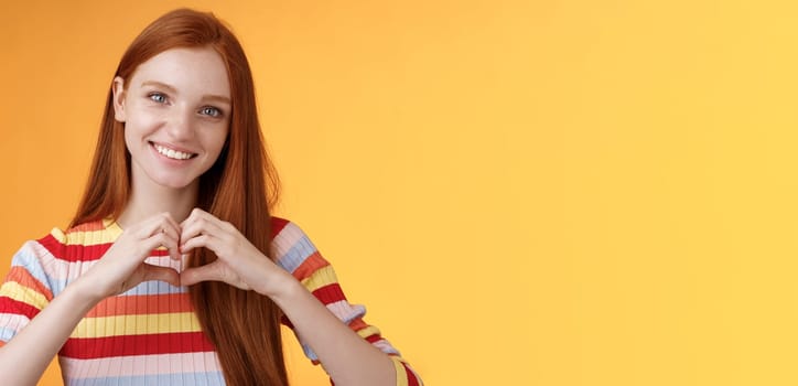 Lovely friendly-looking charismatic smiling redhead girl express affection love and friendship grinning show heart sign chest showing sympathy, confess romantic feelings, standing orange background.