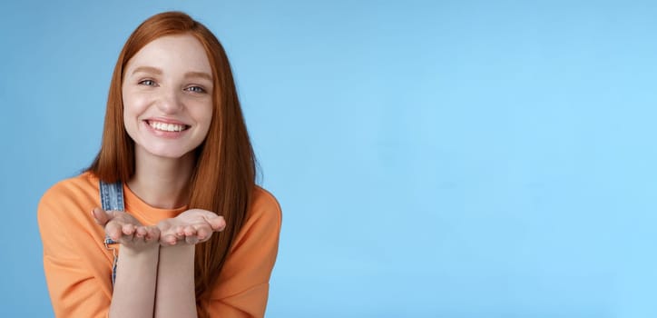 Cute tender kind young ginger girl giving all love you, hold something palms showing camera smiling delighted introduce present grinning romantic gesture send air kisses, blue background.