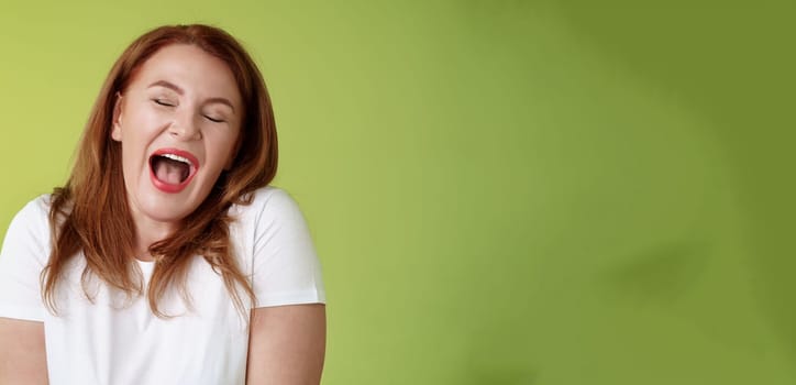 Lazy weekends finally time sleep. Cheerful redhead middle-aged 50s woman yawning satisfied close eyes feel sleepy wake up early morning wanna take nap stand green background watching boring film.