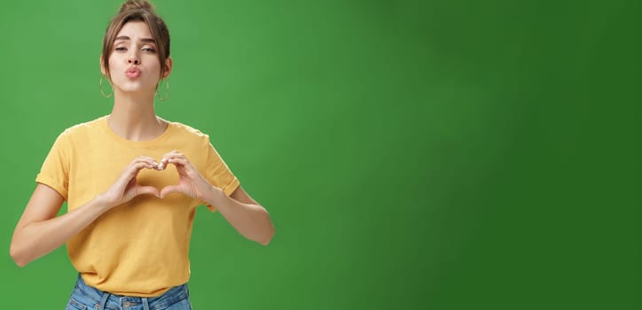 Sensual charming woman in yellow t-shirt and combed hairstyle in yellow t-shirt showing heart gesture against chest folding lips to give kiss or mwah tender and loving over green background. Lifestyle.