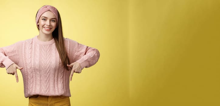 Tender young 20s european girl in sweater smiling broadly finally take-off braces pointing down promoting skillful dentist professional. Female student standing happy positive over yellow wall.
