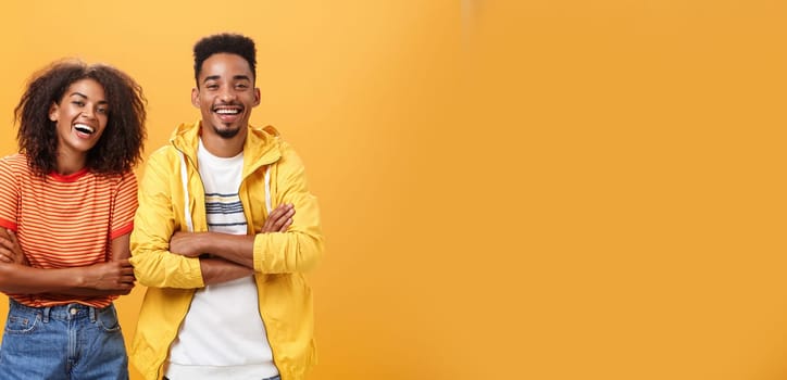 Two african american man and woman being best friends laughing out loud watching funny movie in cinema all dressed up in stylish outfit. standing with hands crossed on chest and amused expression.